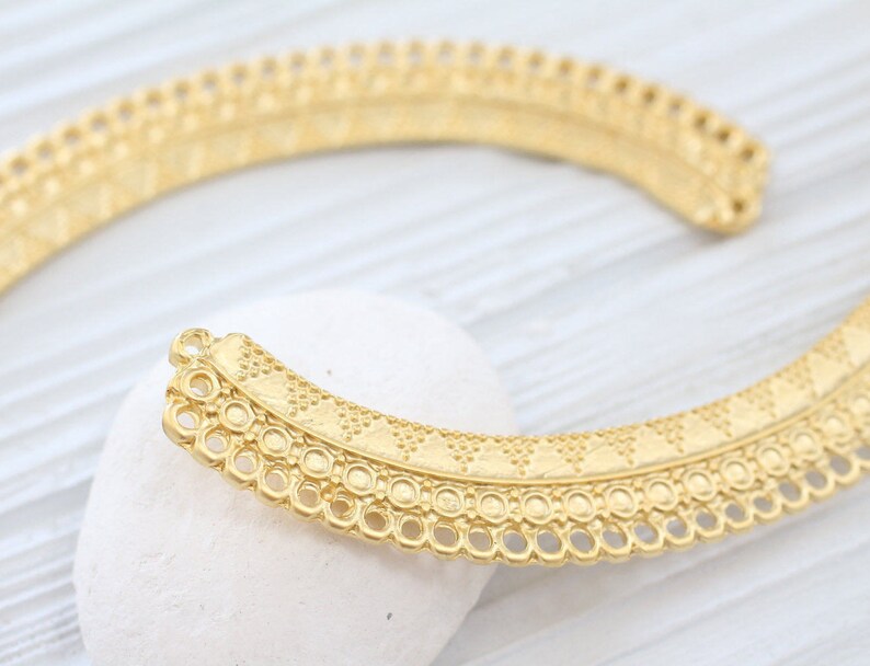 May include: Gold-toned metal jewelry component with a decorative, intricate design. The component is shaped like a half-circle and has a scalloped edge.