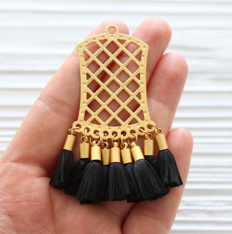 May include: A gold-colored metal pendant with a lattice design and small black tassels. The pendant has a rectangular shape with a rounded top and a small loop for hanging. The tassels are attached to the bottom of the pendant.