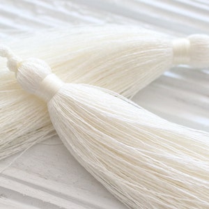 May include: Two white tassel trim pieces with a long, thin fringe. The tassels are made of a soft, silky material and have a delicate, elegant look.