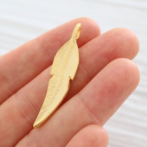 May include: A gold-colored metal feather charm pendant. The feather has a detailed design and a loop at the top for attaching to a necklace or bracelet.
