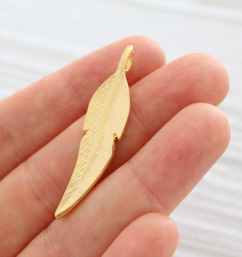May include: A gold-colored metal feather charm pendant. The feather has a detailed design and a loop at the top for attaching to a necklace or bracelet.
