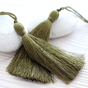 May include: Two olive green tassel trims with long strands of fringe. The tassels are tied together with a thin cord.