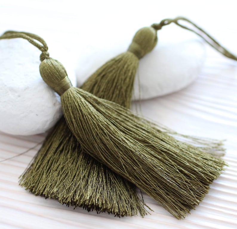 May include: Two olive green tassel trims with long strands of fringe. The tassels are tied together with a thin cord.