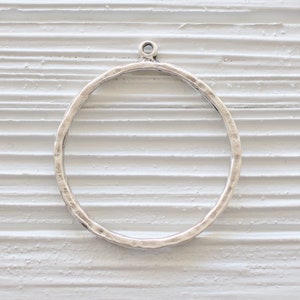 May include: A silver-colored, hammered metal ring pendant with a small loop at the top for attaching to a chain or other jewelry component. The ring has a textured surface and a circular shape.