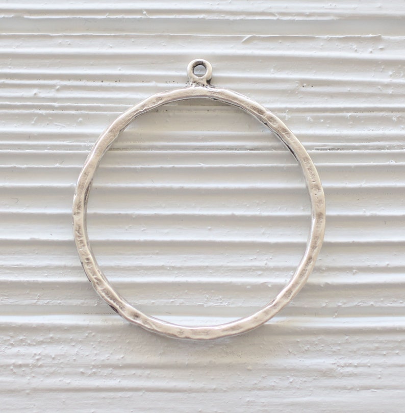 May include: A silver-colored, hammered metal ring pendant with a small loop at the top for attaching to a chain or other jewelry component. The ring has a textured surface and a circular shape.