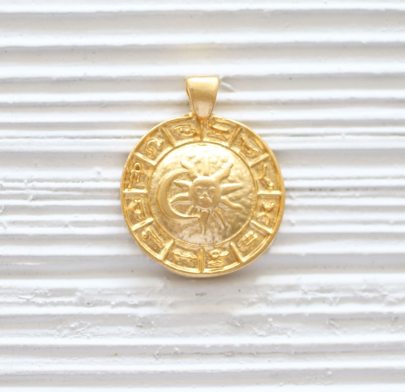 Puede incluir: Colgante dorado con un dise&ntilde;o circular con los signos del zod&iacute;aco y un gr&aacute;fico de sol y luna.
