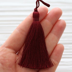 May include: A deep burgundy tassel with a long, flowing fringe. The tassel is held in a hand, showcasing its size and texture.