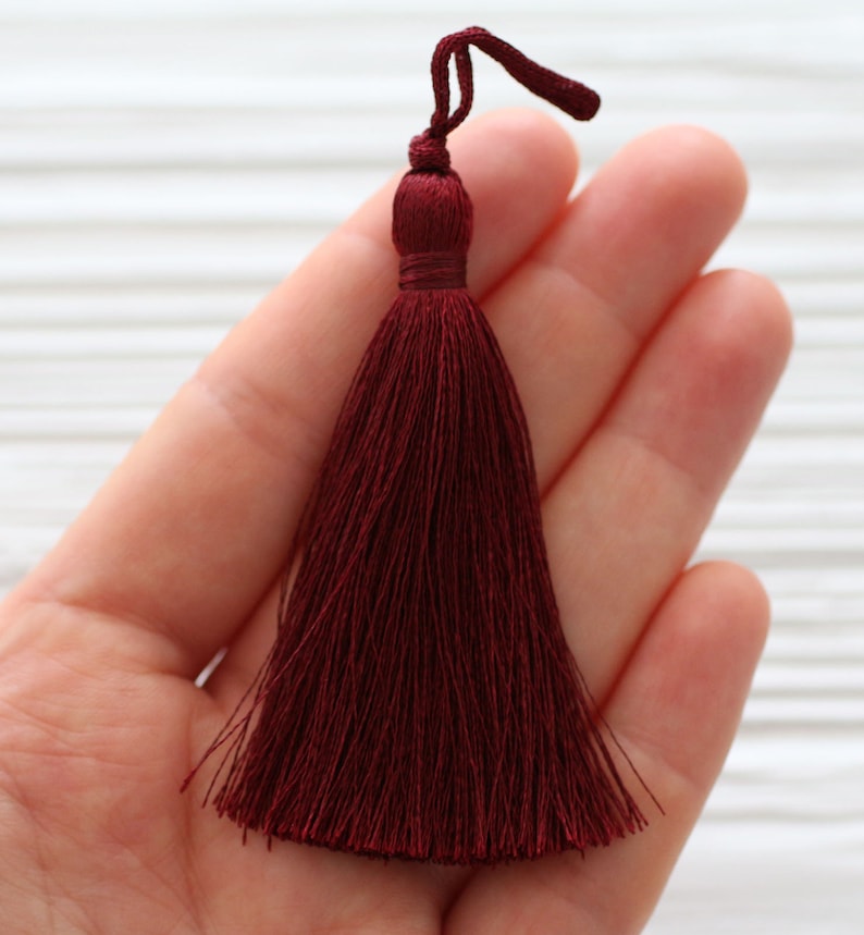 May include: A deep burgundy tassel with a long, flowing fringe. The tassel is held in a hand, showcasing its size and texture.