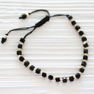 Può includere: Un braccialetto con perline nere e dorate, con un cordoncino nero e chiusura regolabile. Il braccialetto presenta perline quadrate con dettagli dorati.