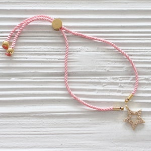 Puede incluir: Un brazalete ajustable de cordón rosa con un dije de estrella dorada. La estrella está incrustada con pequeñas piedras transparentes.