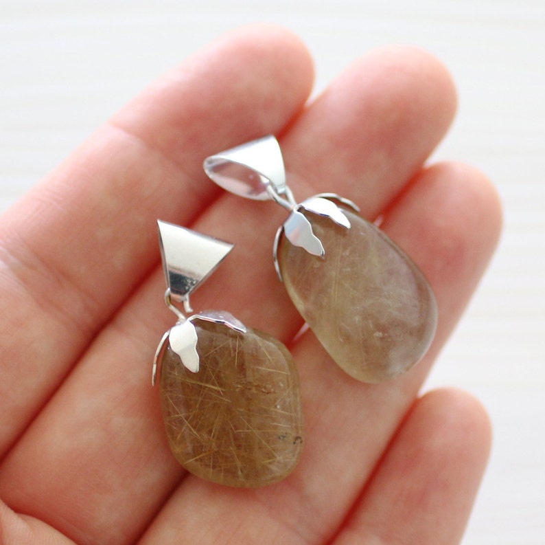 May include: Two gold rutilated quartz pendants with silver bail settings. The pendants are oval shaped and have a translucent, golden color with visible inclusions of rutile.