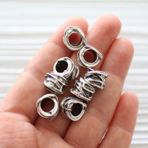 Puede incluir: Cuentas de metal plateado con una superficie texturizada. Las cuentas tienen un agujero en el centro y están diseñadas para ser usadas como ataduras para el cabello o joyas.