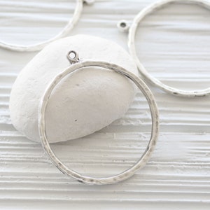 May include: Several silver-colored metal circle pendants with a hammered texture are displayed. Each pendant has a small loop at the top for attaching to a chain or other jewelry component. The pendants are on a white background.