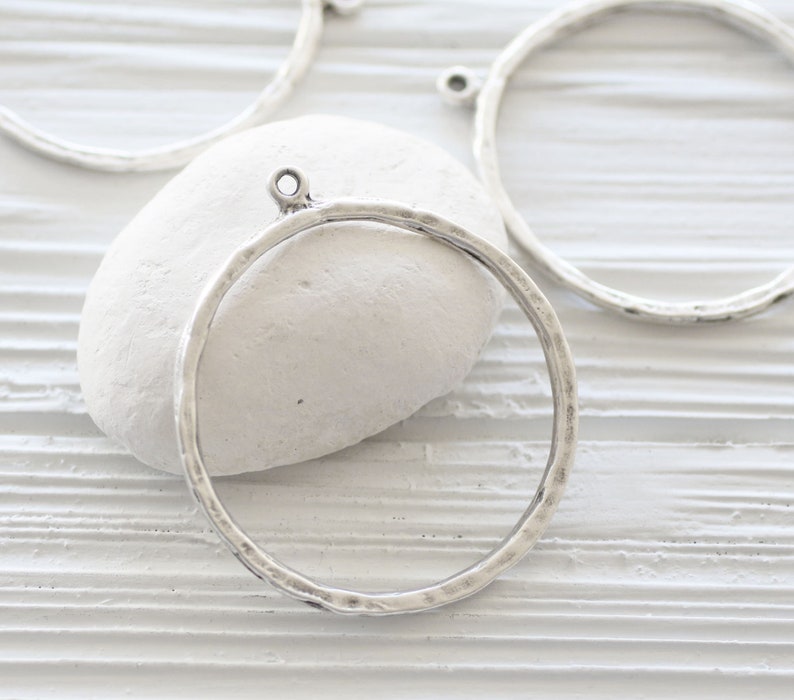 May include: Several silver-colored metal circle pendants with a hammered texture are displayed. Each pendant has a small loop at the top for attaching to a chain or other jewelry component. The pendants are on a white background.