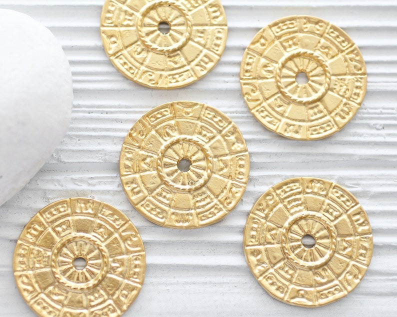 Pu&ograve; includere: Cinque ciondoli rotondi in metallo color oro con disegni intricati. Ogni ciondolo presenta un motivo circolare con un foro centrale. I ciondoli hanno un bordo leggermente rialzato e una superficie testurizzata.