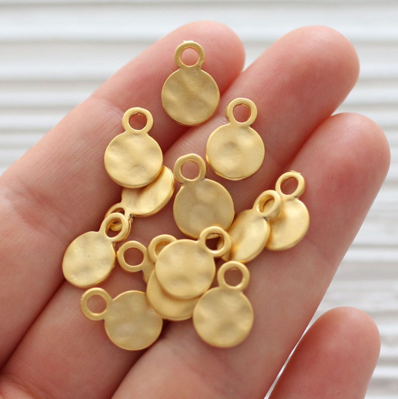 May include: A collection of ten gold-toned metal circle charms with a hammered texture. Each charm has a small loop at the top for attaching to jewelry or other crafts.