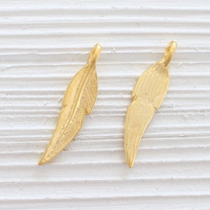 May include: Two gold-colored feather charms with a textured surface. The charms have a loop at the top for attaching to jewelry.