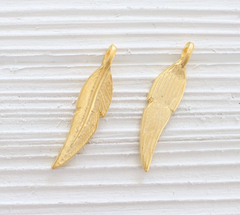 May include: Two gold-colored feather charms with a textured surface. The charms have a loop at the top for attaching to jewelry.