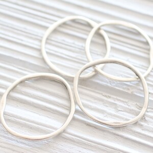 May include: Four silver rings, each with a slightly irregular, rounded shape, are stacked on a white wooden surface.