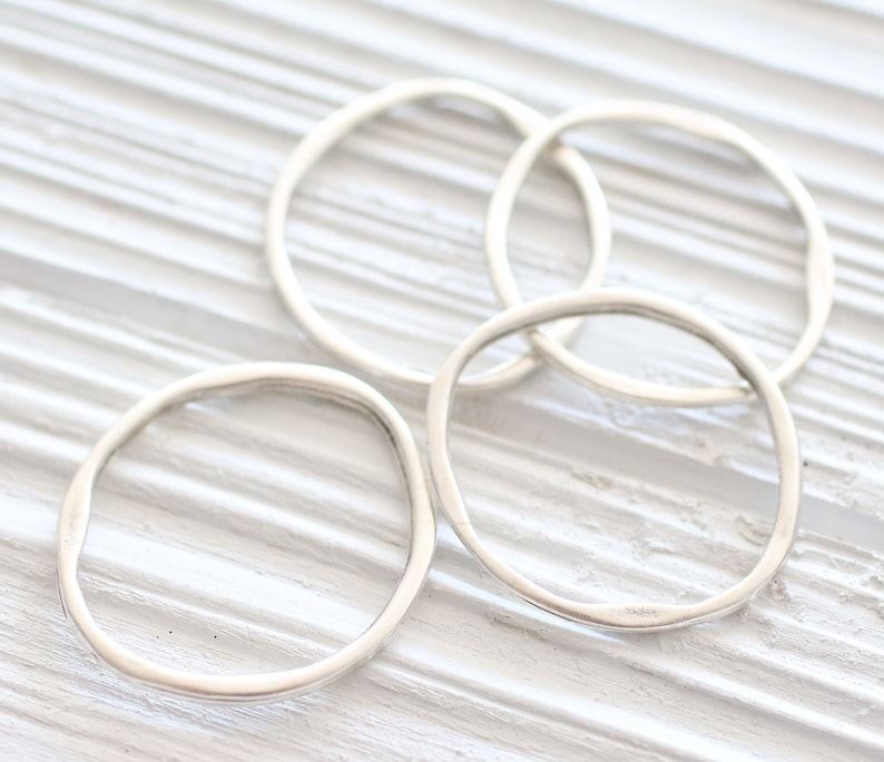 May include: Four silver rings, each with a slightly irregular, rounded shape, are stacked on a white wooden surface.