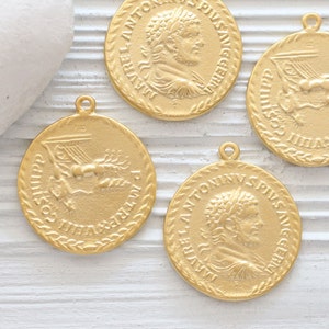 May include: Four gold-colored metal charms with Roman-inspired designs. Each charm features a different image, including a profile of a man with a laurel wreath, a winged figure, and a lyre. The charms have a rope-like border.