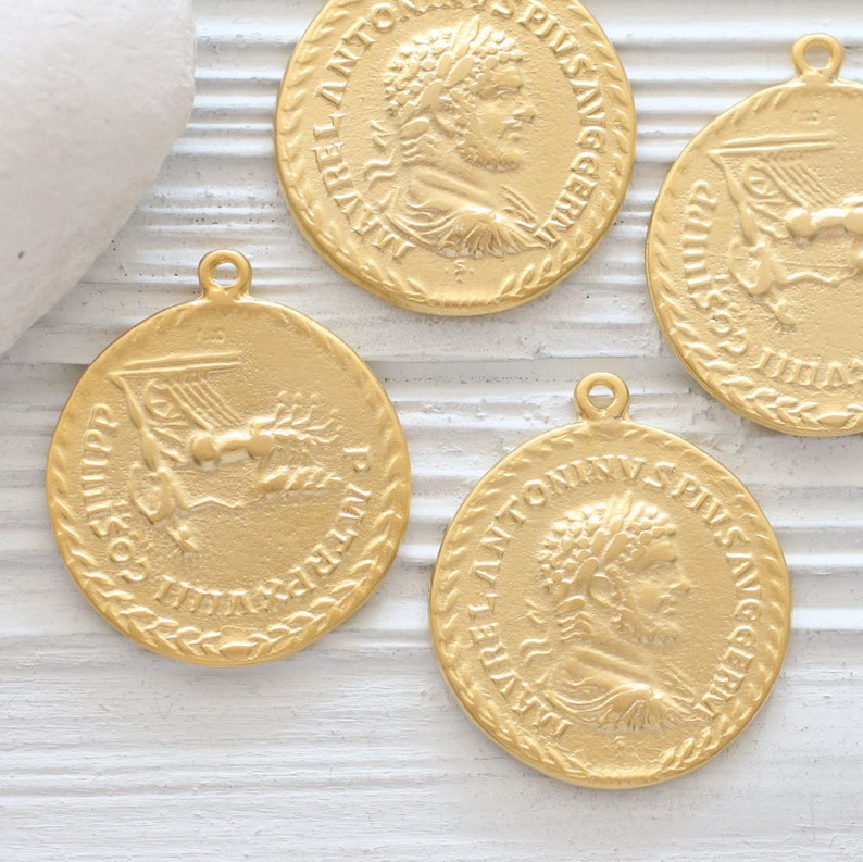 May include: Four gold-colored metal charms with Roman-inspired designs. Each charm features a different image, including a profile of a man with a laurel wreath, a winged figure, and a lyre. The charms have a rope-like border.