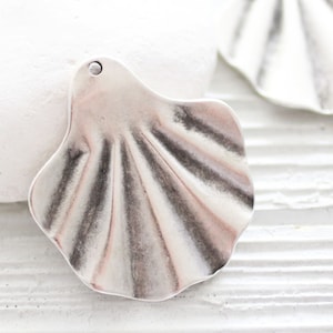 May include: A silver-toned metal seashell charm with a textured surface. The charm has a hole at the top for attaching to jewelry or other crafts.