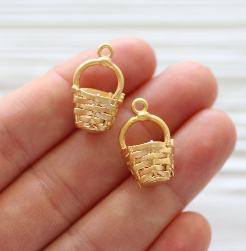 May include: Two gold-colored metal basket charms with a woven design. The charms have a loop at the top for attaching to jewelry or other crafts.