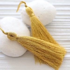 May include: Two metallic gold tassels featuring a tightly wrapped cord head and a long, flowing fringe of fine, shimmering threads. Each tassel includes a small loop at the top for attachment, showcasing a bright, reflective finish throughout.