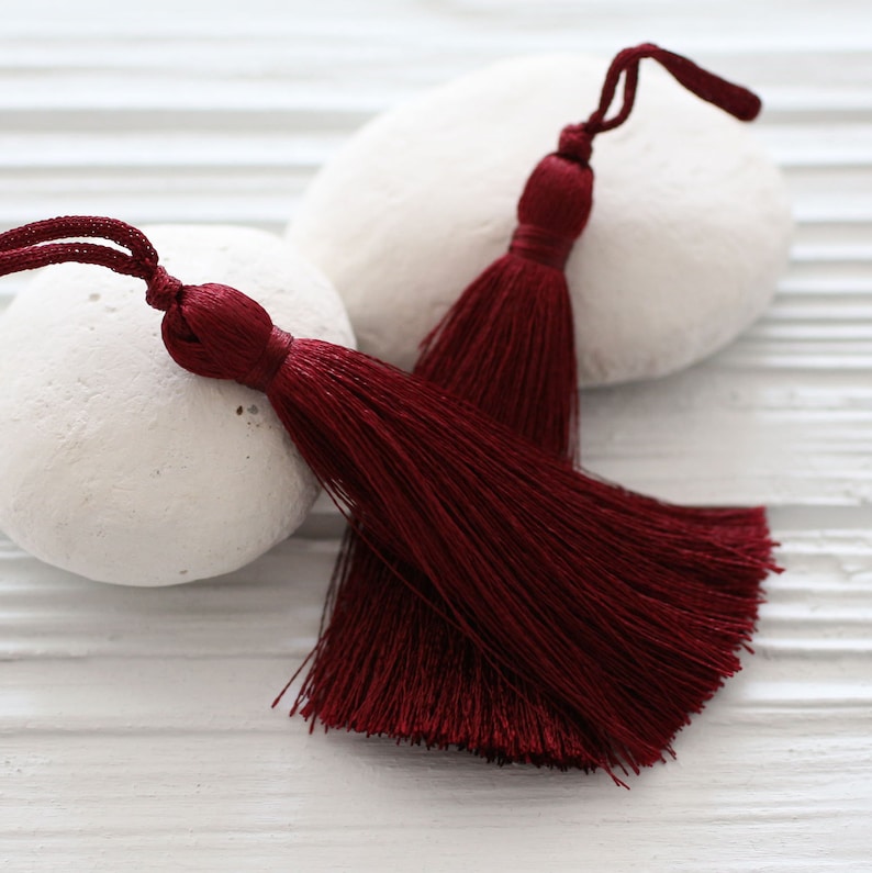 May include: Two burgundy silk tassels with a long fringe. The tassels are on a white background.