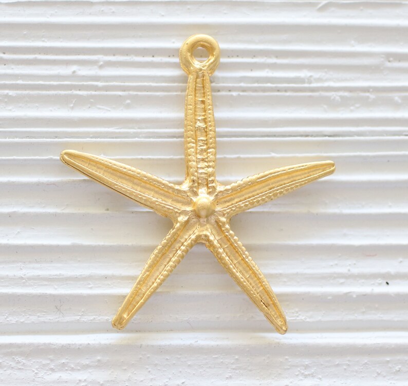 May include: Gold-toned metal starfish charm with a loop at the top for hanging. The starfish has a detailed, textured surface.