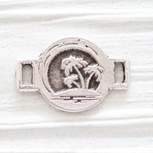 May include: Silver charm with a palm tree design. The charm has two loops on the sides for attaching to a bracelet or necklace.