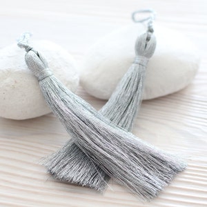 May include: Two silver tassel trim with a metallic sheen. The tassels are on a white surface.