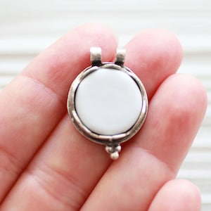 May include: A silver pendant with a white round ceramic insert. The pendant has a loop at the top for attaching to a chain.