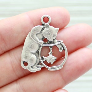 May include: Silver charm of a cat looking into a fishbowl with a fish inside. The charm has a loop at the top for attaching to a necklace or bracelet.
