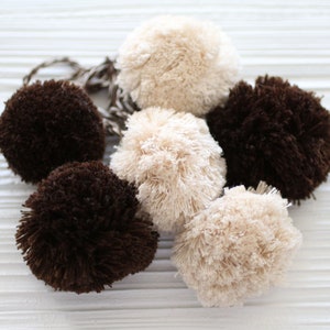 May include: A selection of fluffy pom-poms in shades of brown and cream. The pom-poms are arranged in a cluster, with a few connected by a brown and white twisted cord. The texture of the pom-poms appears soft and plush.