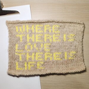 以下が含まれることがあります： ベージュ色のニットスクエアで、明るい黄色の文字で「WHERE THERE IS LOVE THERE IS LIFE」と書かれています。 ニット作品は、白い紙の隣の木製の表面に置かれています。