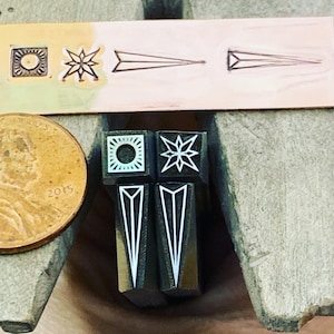 Puede incluir: Cuatro sellos de metal con diseños geométricos. Los sellos son negros con diseños blancos. Los diseños incluyen un sol, una estrella y dos triángulos.