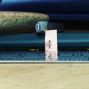 May include: A small, rectangular piece of metal with a tree design stamped into it. The metal is a light rose gold color.