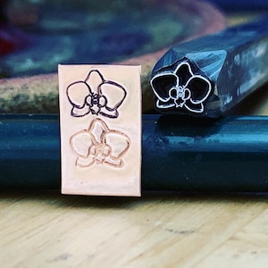 May include: A close-up of a metal stamp with an orchid design. The stamp is used to create impressions on metal. The stamp is shown next to a piece of metal with two orchid impressions.
