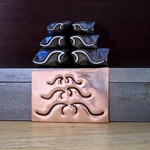 May include: A collection of metal stamps with a wave design. The stamps are dark gray with white accents and are stacked on a wooden surface. A copper-colored stamp with the wave design is in the foreground.