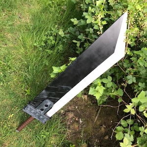 May include: A large, black and white sword prop with a brown handle. The blade is primarily black with a white edge, and the hilt features grey metal detailing. The sword is lying on a bed of green grass and foliage.