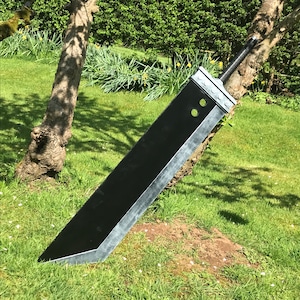 May include: A large black and silver sword rests against a tree on grass. The sword features a rectangular blade with a silver edge and a black handle. Two circular holes are visible near the hilt.
