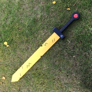 May include: A yellow toy sword with a black handle and guard, and a red circle at the pommel. The blade has black markings and is lying on green grass.