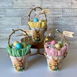 May include: Three small decorative Easter baskets with bunny designs. Each basket is filled with pastel colored tissue paper and plastic eggs. The baskets are decorated with the text "Happy Easter".