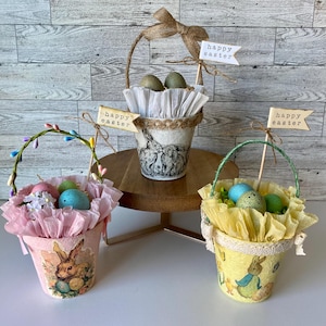 May include: Three small Easter baskets with pastel pink, yellow, and white tissue paper lining. Each basket has a different design and contains plastic Easter eggs. The baskets are decorated with bunny and floral designs. The baskets have signs that say "Happy Easter".