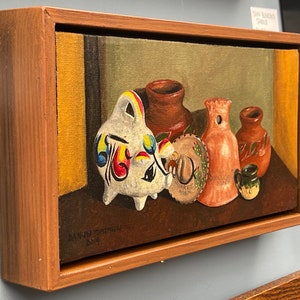 May include: A still life painting of colorful pottery, including a white piggy bank with red, yellow, blue, and green accents, a brown clay pot, a pink clay pot with a hole in the top, a brown clay pot with a green design, and a small brown clay cup. The painting is framed in a wooden frame.