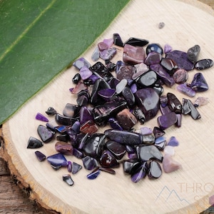 May include: A collection of small, purple and black amethyst chips on a wooden surface. The chips are various shapes and sizes, and some have a shimmery finish.