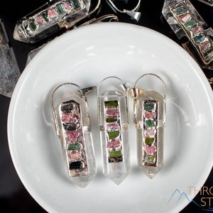 This crystal pendant features a clear Quartz gemstone, showcasing its transparent beauty and amplifying properties. Surrounding the Quartz are seven, green and pink Tourmaline gemstone accent crystals.