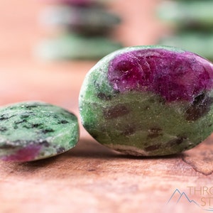 Może przedstawiać: Dwa zielone i różowe zoisity w kształcie kamieni szlifowanych na drewnianej powierzchni. Większy kamień ma wyraźny różowy fragment, podczas gdy mniejszy kamień ma bardziej subtelny różowy odcień.